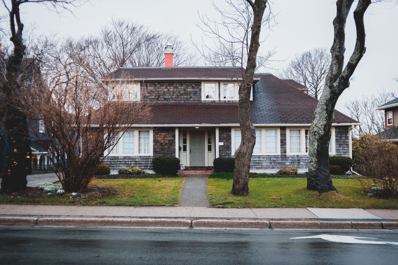 about-04 Facade of modern residential cottage with brown tiled roof surrounded by leafless trees located near road in city in cloudy day